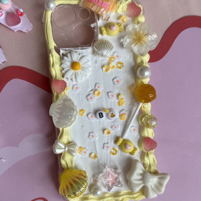 A finished decoden phone case in white and lemon cream with daisies and a lollipop